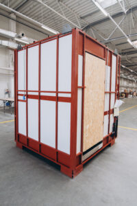 Prefabricated bathroom pod with an OSB door on a red steel frame, in the hall.