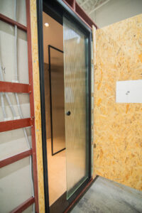 Entrance to a prefabricated bathroom pod — a glass door and shower partition seen from the OSB side.