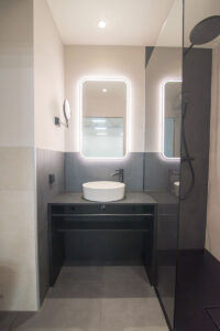 Grey bathroom with backlit mirrors, a countertop basin on a black vanity and a walk-in shower.